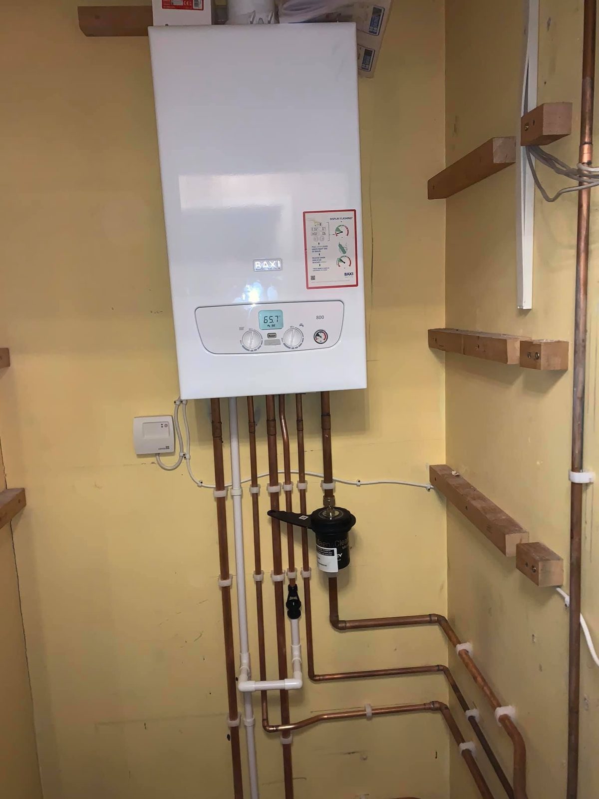 Boiler in a cupboard with pipework and a filter, shelving battens on the wall
